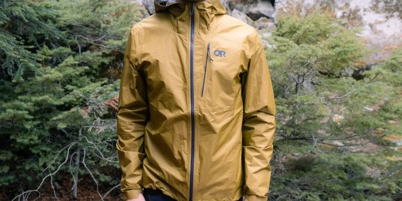 Man wearing a golden ultralight waterproof jacket with an OR logo, standing in a forest.