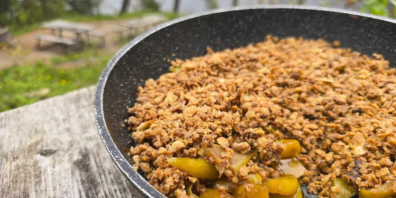 A warm, bubbling campfire apple crisp in a pan, ready to be enjoyed outdoors by a lake.