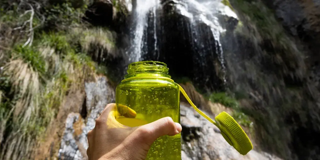 A hand holds a bright green water bottle, ready for a refreshing drink after a long hike. Behind it, a beautiful waterfall cascades down a rocky cliff, surrounded by lush greenery.