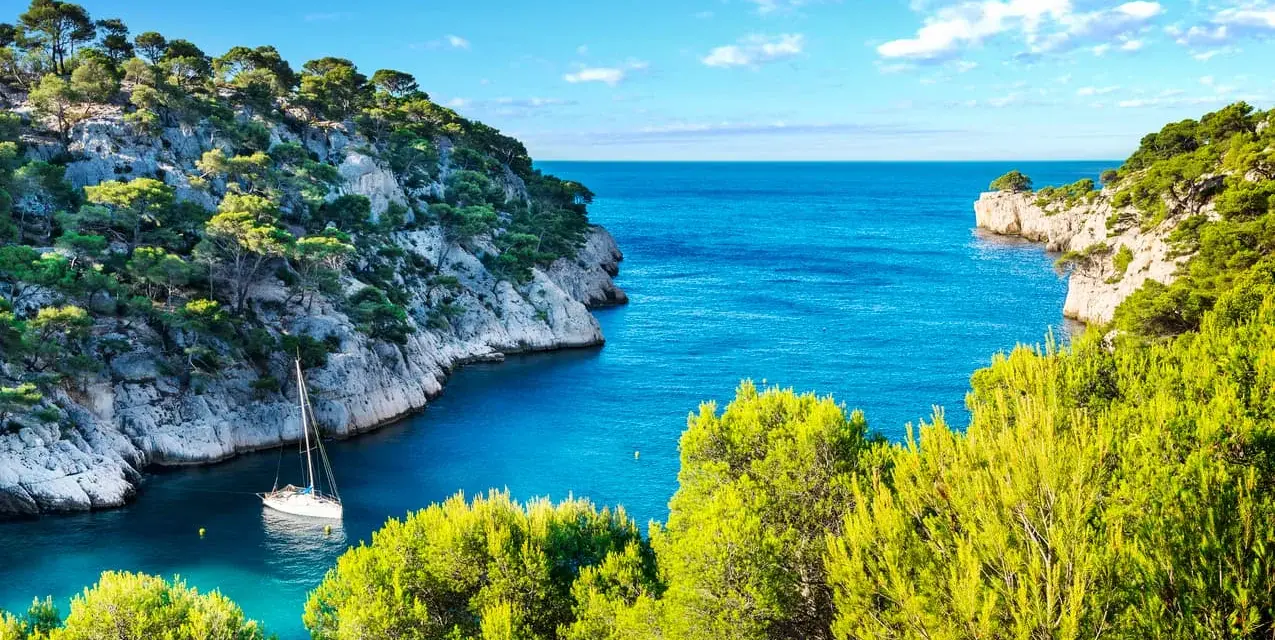 A sailboat anchors in a serene cove, surrounded by lush green trees and dramatic cliffs. This idyllic scene evokes the feeling of a perfect camping trip in Cassis.