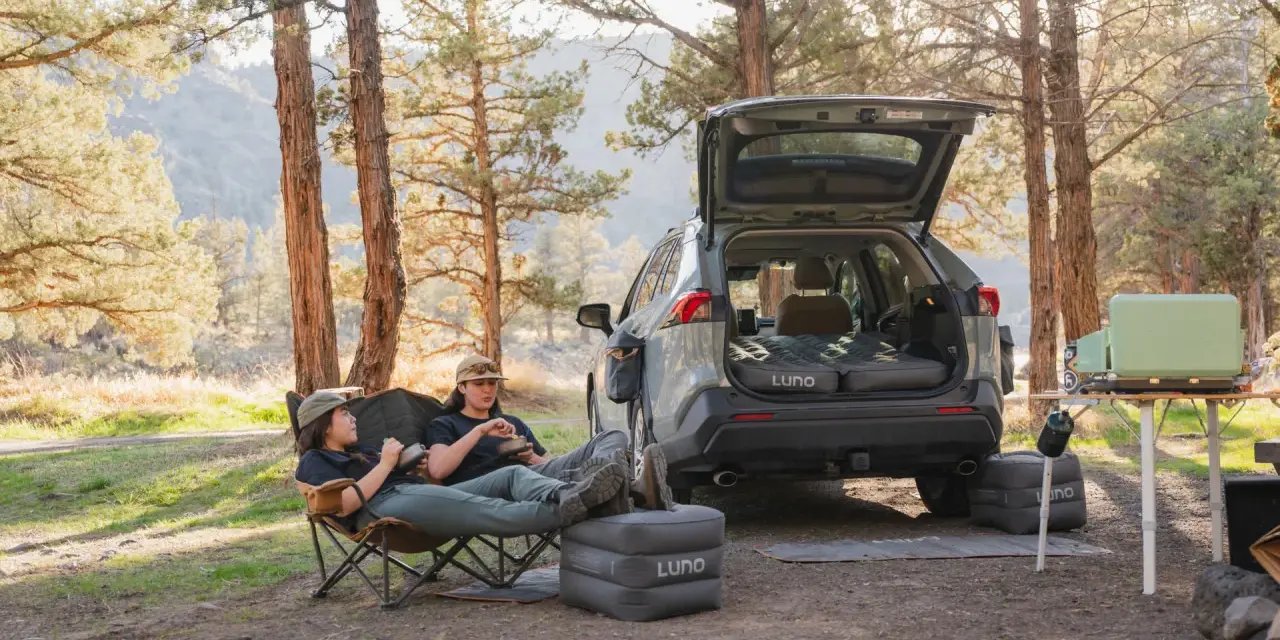 Two people enjoy a meal in camping chairs next to a car with an open trunk, showcasing clever car camping hacks with an inflatable mattress and a portable stove.