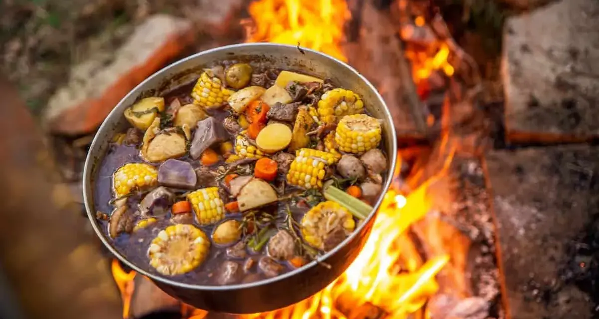 A pot of hearty camping meals, filled with corn, potatoes, carrots, and meat, simmers over a crackling campfire.