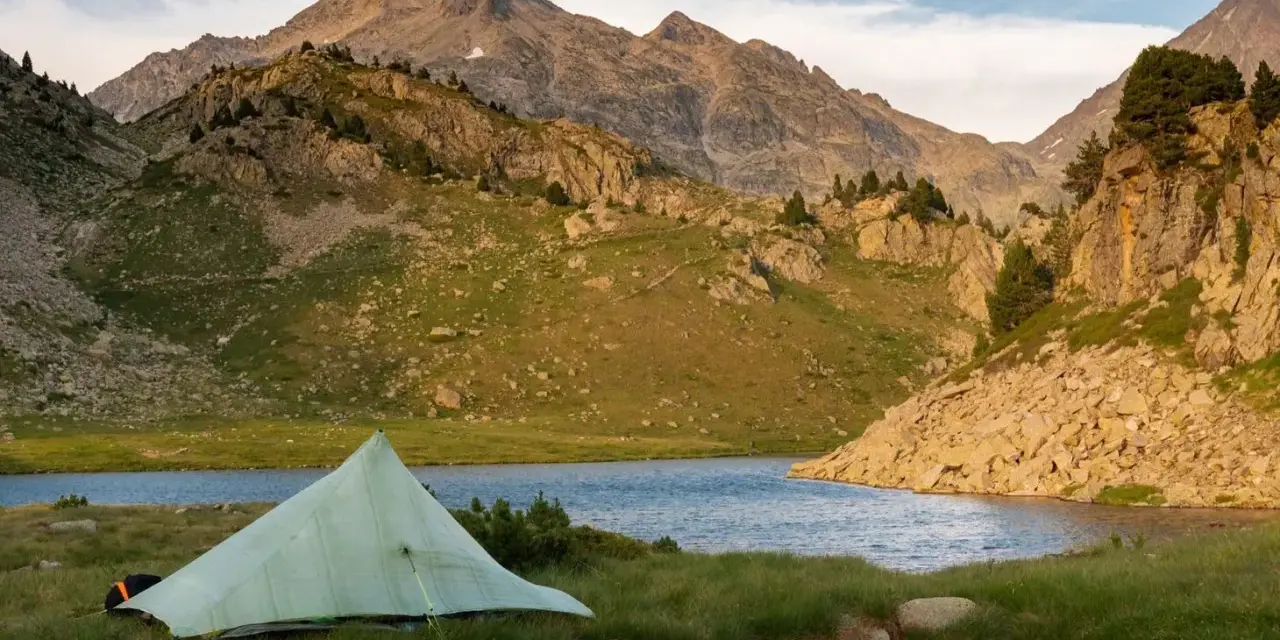 A tent sits by a serene lake, with rugged mountains in the background, a perfect campsite on the Pyrenees High Route.
