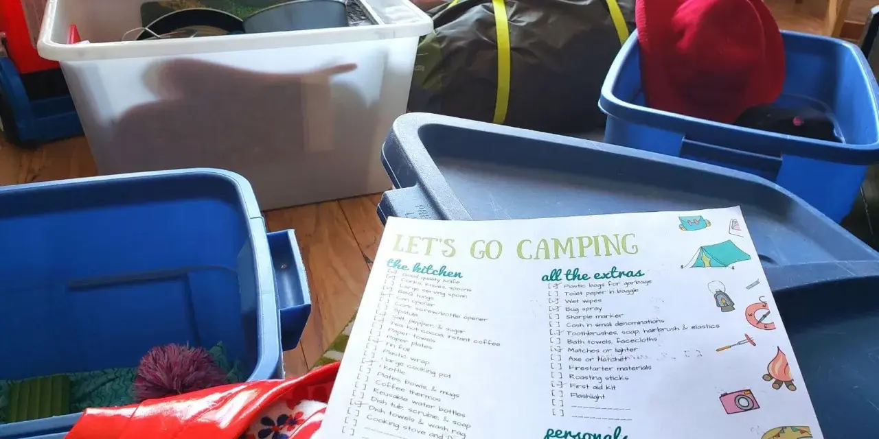 A "Let's Go Camping" checklist is spread out among blue and white storage bins, ready for a trip.