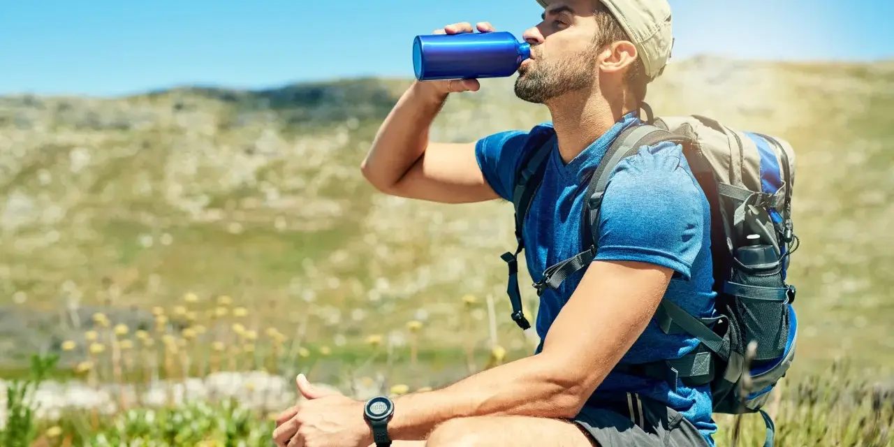 A hiker takes a refreshing drink from a blue water bottle, demonstrating how to stay hydrated while hiking in a sunny, mountainous landscape.
