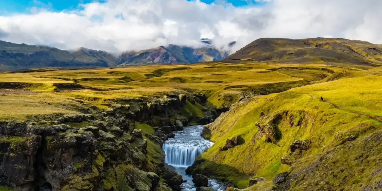A winding path on the Laugavegur trail in Iceland leads past a mossy canyon with a cascading waterfall.