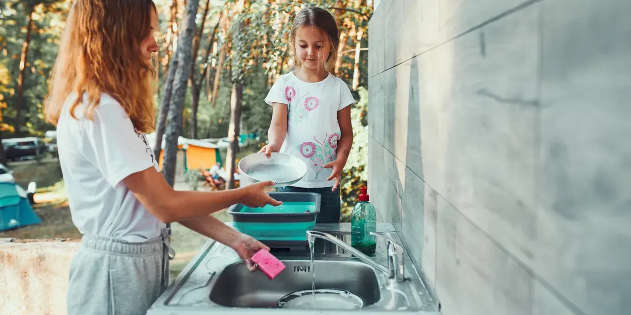 Two girls are washing dishes camping. One holds a pan over a sink while the other prepares to rinse it.