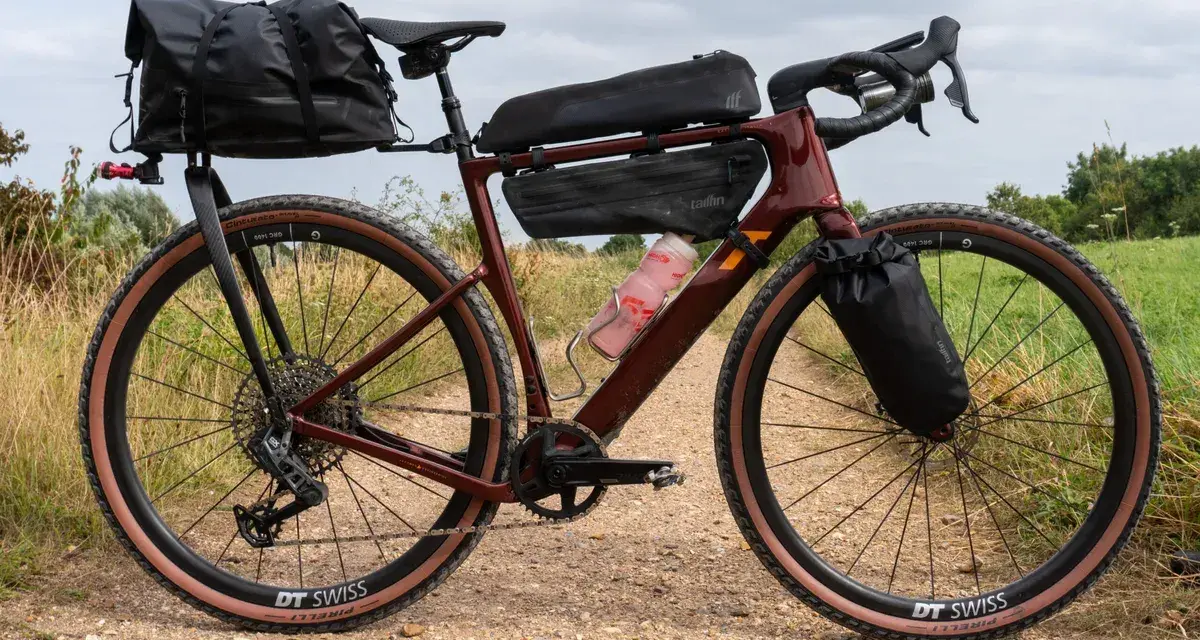 A maroon gravel bike with a full bikepacking setup, including saddle and frame bags, ready for an adventure on a dirt path.