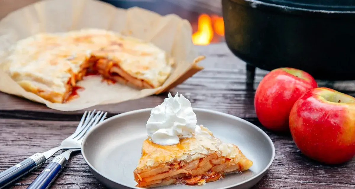 A slice of campfire apple pie, topped with whipped cream, sits on a plate next to fresh apples and a fork.