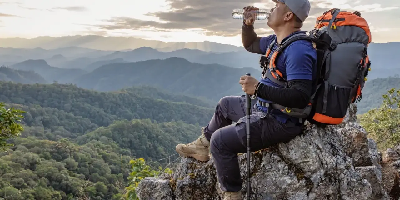 A hiker drinks water on a mountain peak, contemplating how much water to take backpacking.