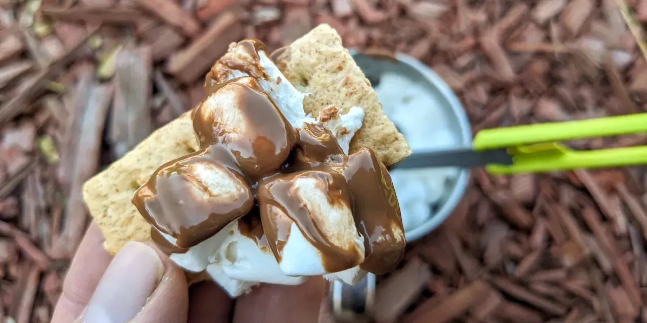 A hand holds a graham cracker topped with melted marshmallows and chocolate, ready to be assembled into a s'more. A pan of marshmallows is visible in the background.