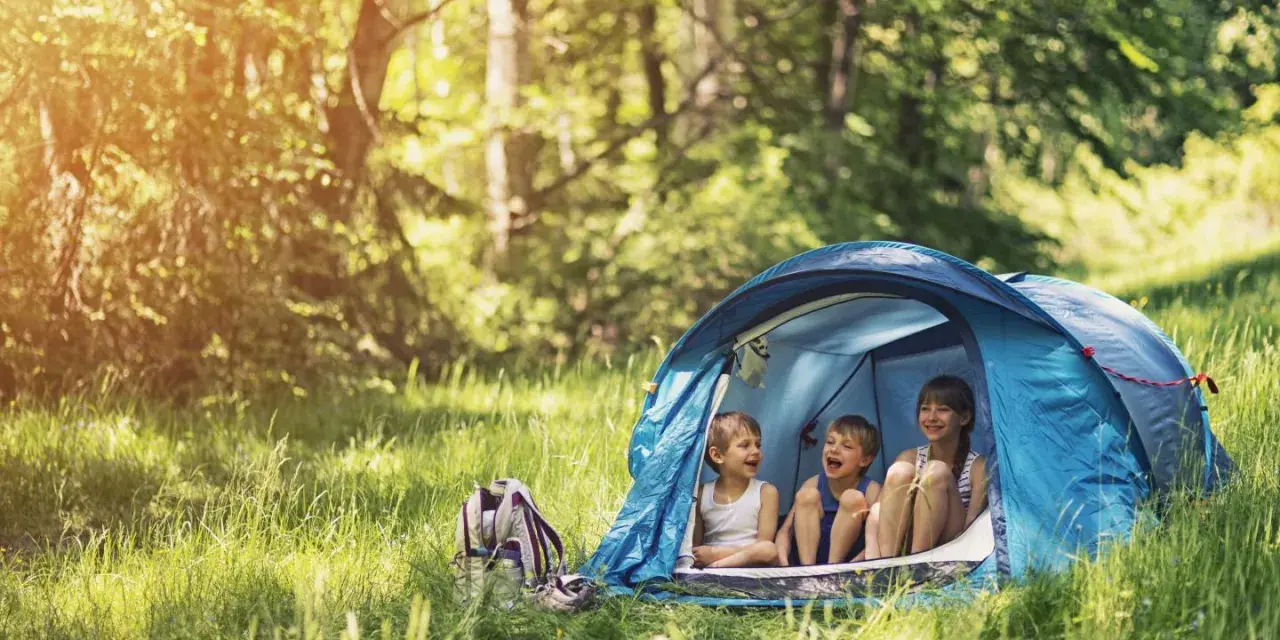Three children laugh inside a blue tent, enjoying camping activities in a sun-dappled forest.