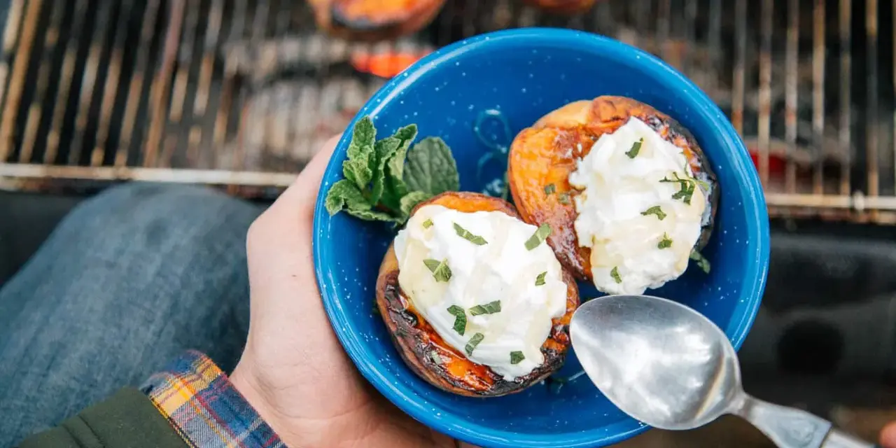 A hand holds a blue bowl with two grilled peaches topped with cream and mint. Campfire peaches are a perfect dessert.