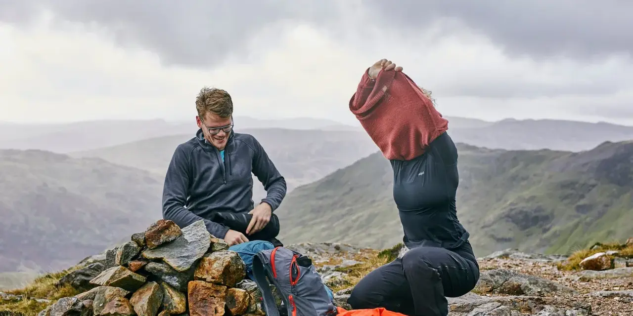 Hikers on a rocky summit adjust their backpacking clothes. One man smiles while sitting on a cairn, next to backpacks. Another person pulls on a red sweater.