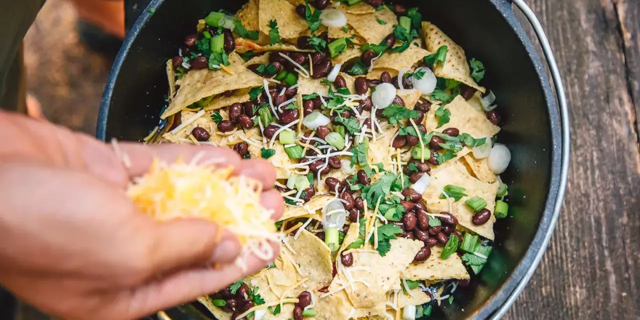Making delicious camping nachos in a Dutch oven, adding shredded cheese to tortilla chips, black beans, and green onions.