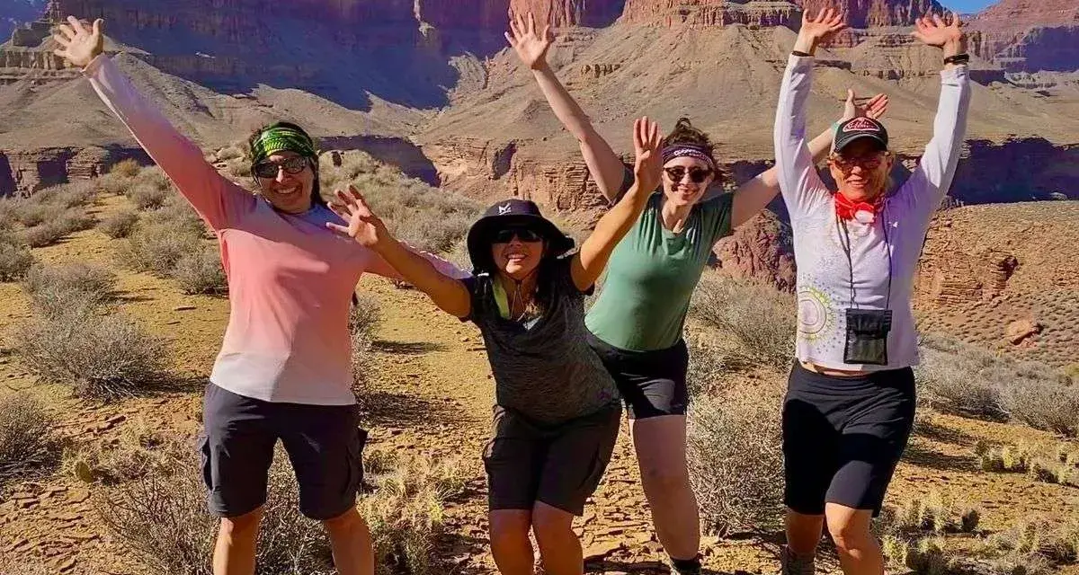 Four women pose with arms raised in a desert canyon, dressed for hiking in hot weather with shorts and light tops.