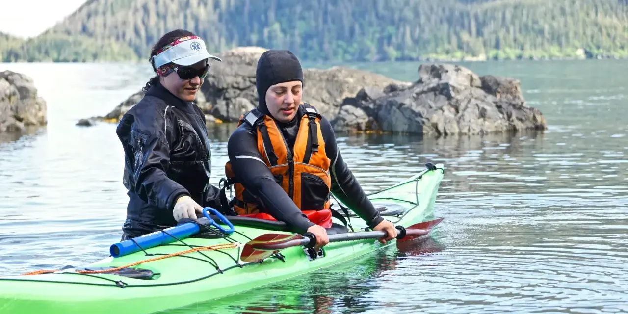 Two kayakers in a bright green kayak practice a paddle float re-entry in calm water, with a forested mountain in the background.