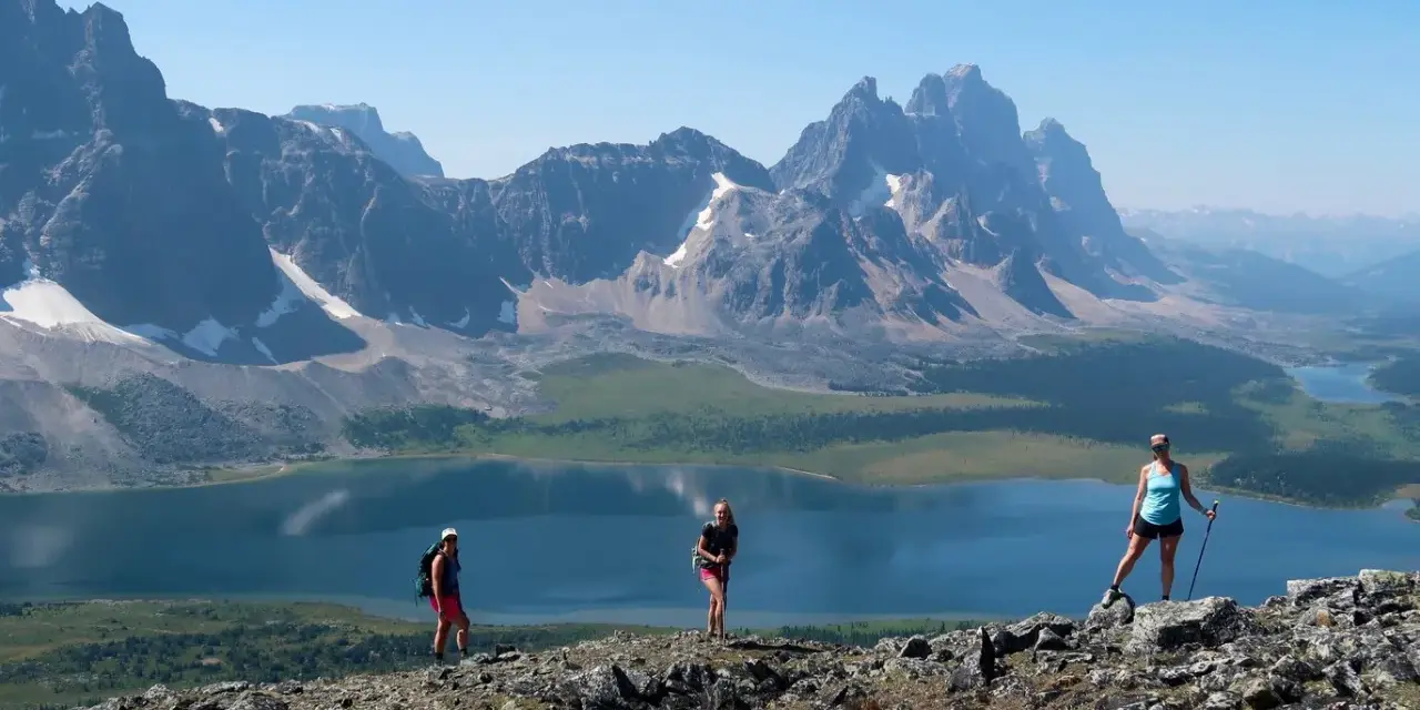 Three hikers enjoy a breathtaking view during their Tonquin Valley hike, with a stunning blue lake and jagged mountains behind them.