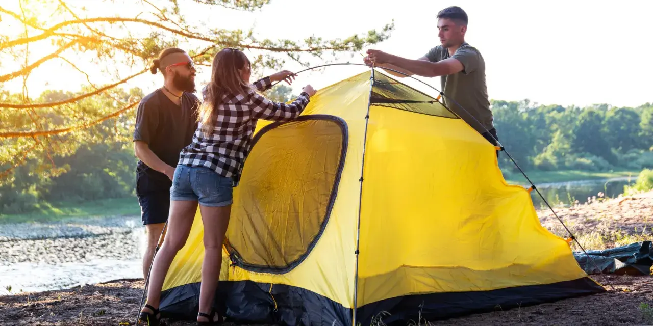Friends setting up a bright yellow tent by a lake, learning how to choose a campsite with a beautiful sunset.
