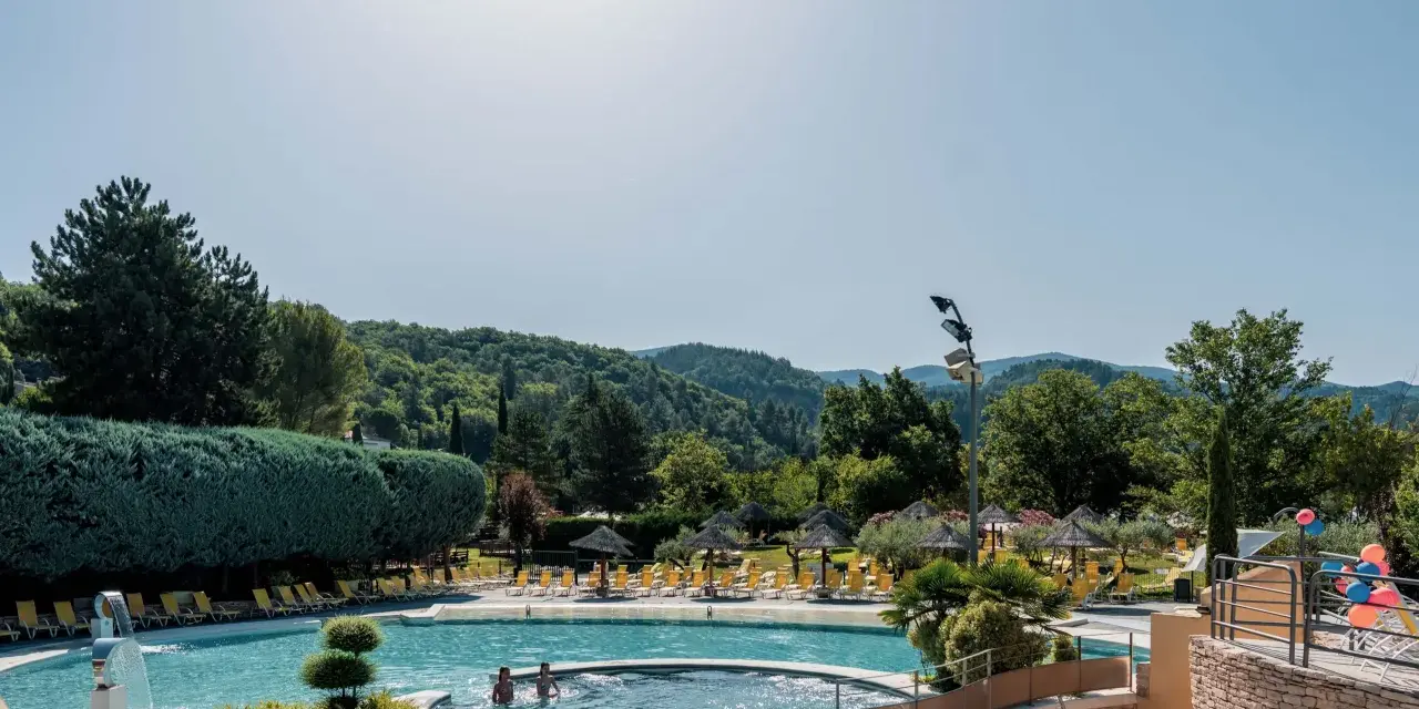 Enjoying a refreshing dip in the pool during a camping trip in Provence. Lush green hills and trees surround the sparkling water, with sun loungers and umbrellas ready for relaxation.
