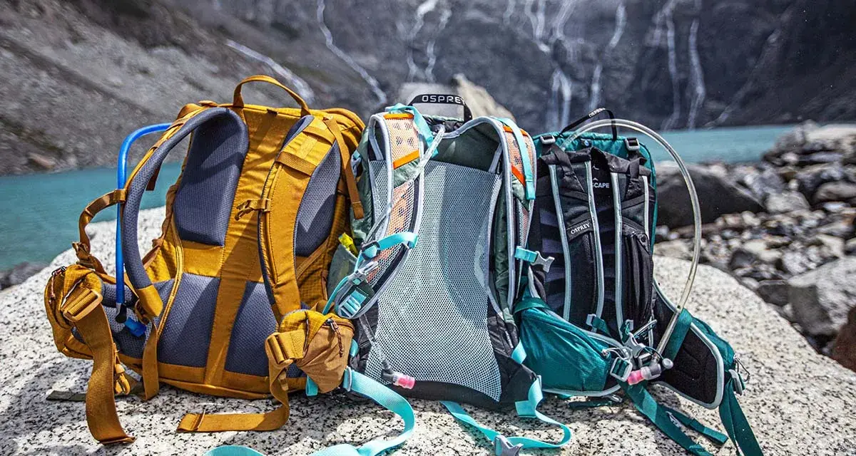 Three Osprey backpacks sit on rocks by a lake, ready for a hike. Learn how to pack a day pack for hiking with these essential tips.
