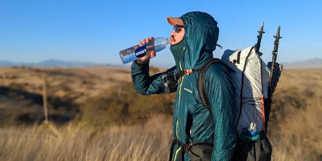A hiker takes a refreshing drink from a Smartwater bottle while carrying a large backpack and trekking poles.