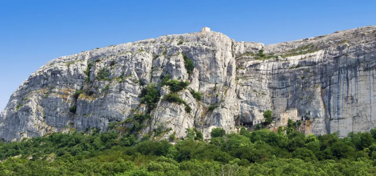 A majestic cliff face rises above a lush green forest, with a small building nestled into the rock at Sainte Baume.