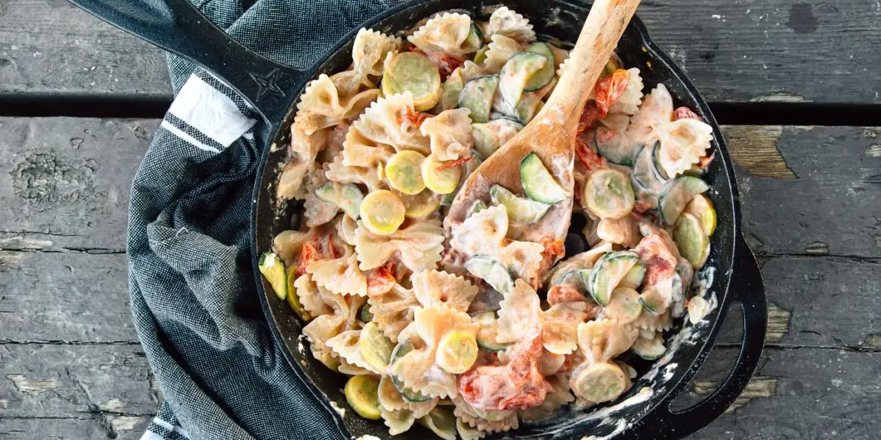 A hearty camping pasta dish with farfalle, zucchini, and tomatoes in a creamy sauce, served in a cast-iron skillet.