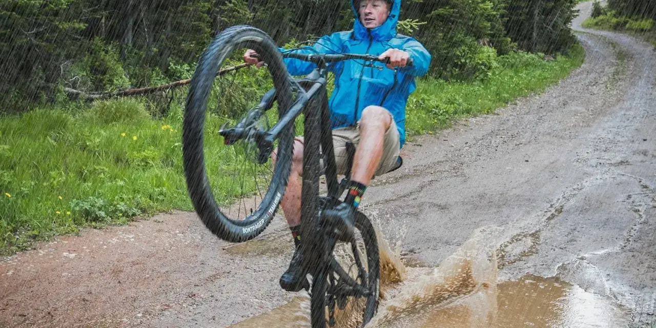A mountain biker in a blue jacket performs a wheelie through a large puddle, splashing water as it rains heavily on a forest trail.