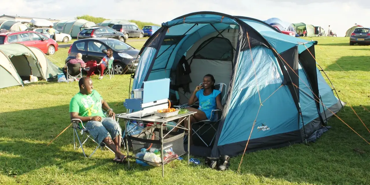 A couple enjoys a meal at their campsite, a perfect example of how to camp. They sit in chairs beside a blue tent, with food laid out on a portable table.