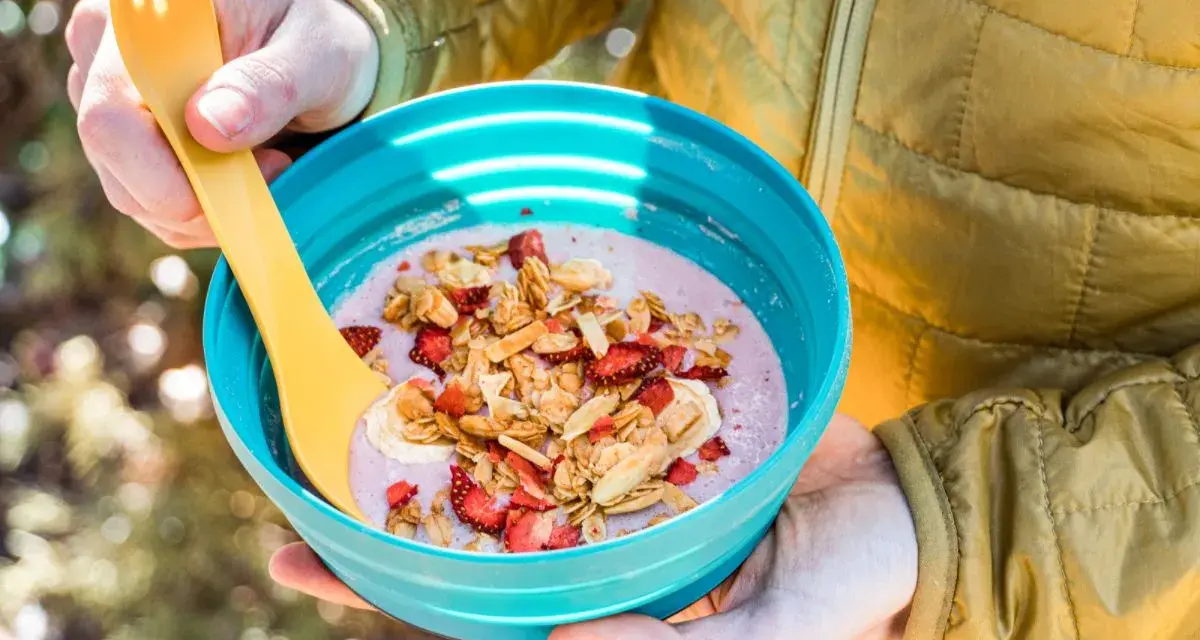 A person in a yellow jacket holds a blue bowl filled with a pink smoothie topped with granola, dried strawberries, and banana slices. Perfect for camping breakfast ideas.