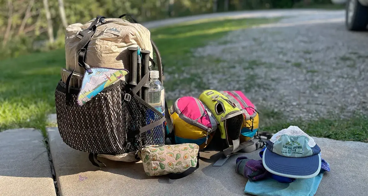 Ready for adventure! A large backpack, hiking poles, water bottle, and colorful pouches are laid out with camping gear, including a hat and gloves.