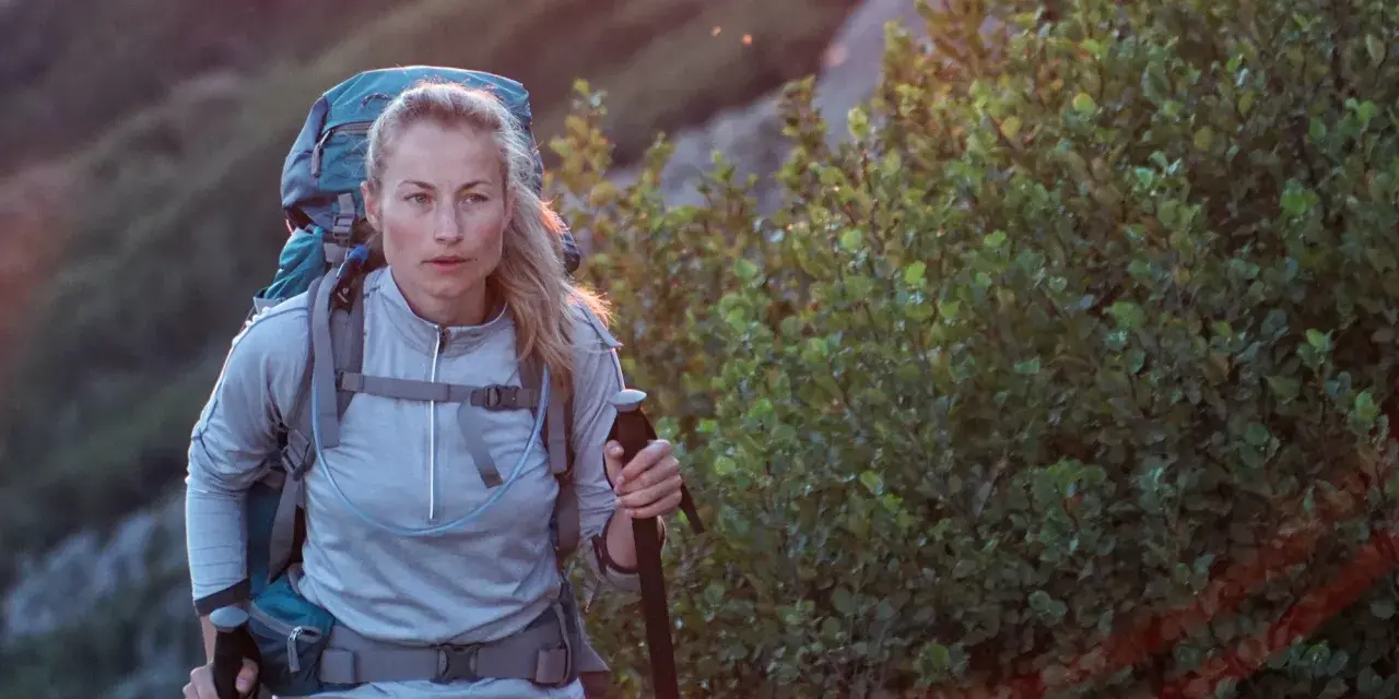 A woman with blonde hair, wearing a grey jacket and a large backpack, is backpacking hiking up a rocky, vegetated slope. She holds a trekking pole.