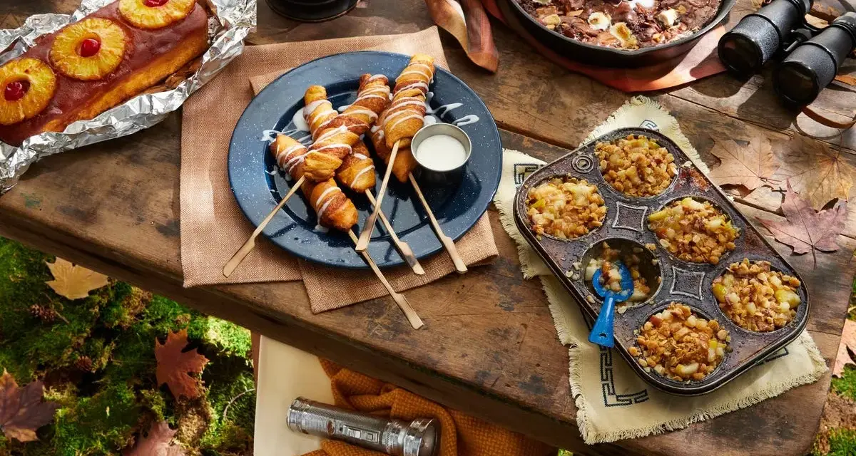 A spread of cozy camping food: pineapple upside-down cake, pigs in a blanket, apple crumble muffins, and a skillet brownie with ice cream.