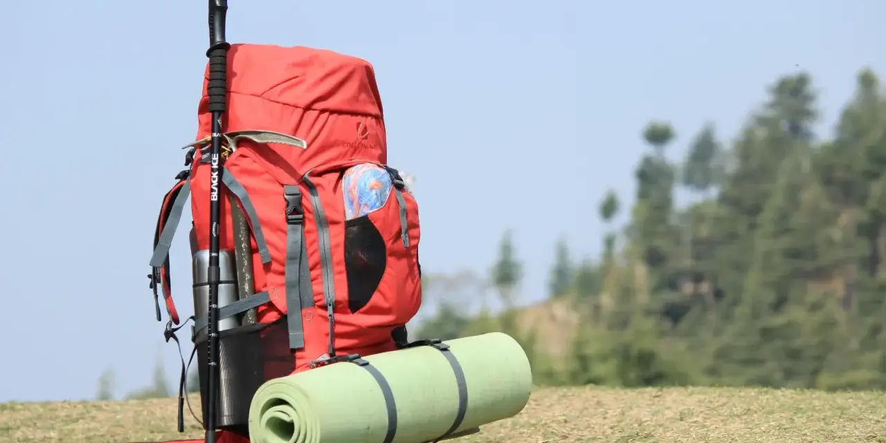 A red backpack with a rolled sleeping pad strapped to it, and trekking poles. This image shows how to pack a backpacking pack for an adventure.