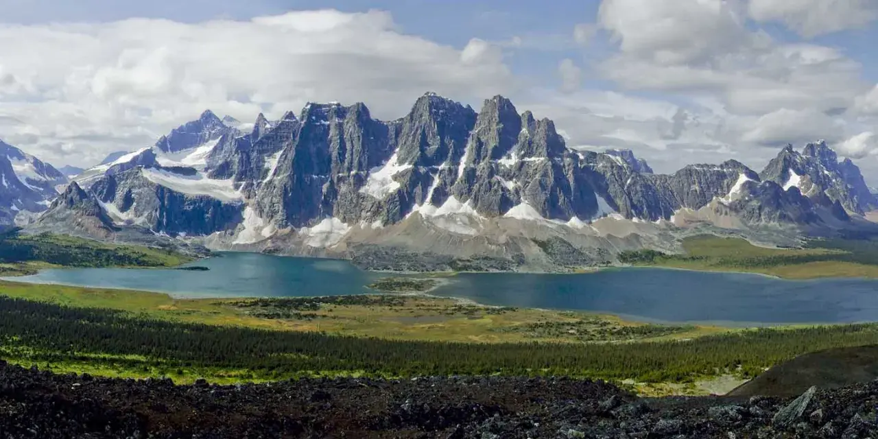 A vast mountain range with snow-capped peaks overlooks a serene blue lake. The Tonquin Valley Trail offers this breathtaking view of rugged beauty.