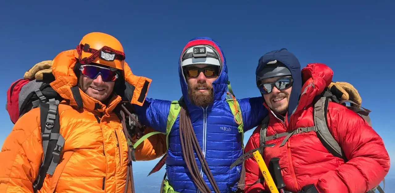 Three smiling mountaineers in bright down fill jackets and helmets pose on a sunny summit, ready for adventure.