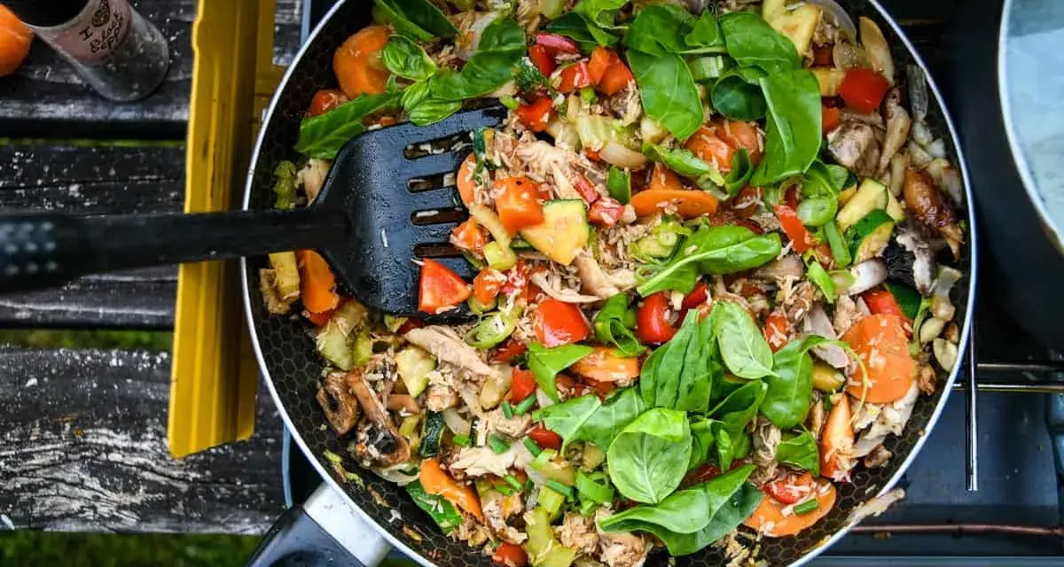 A colorful stir-fry with chicken, carrots, zucchini, and peppers, perfect for camp stove recipes. A black spatula is mixing the ingredients in a pan.