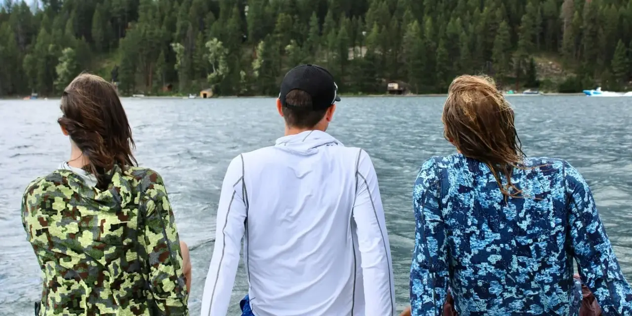 Three friends enjoy a lake view, wearing cooling clothing for extreme heat. The camouflage and blue patterned shirts offer sun protection.