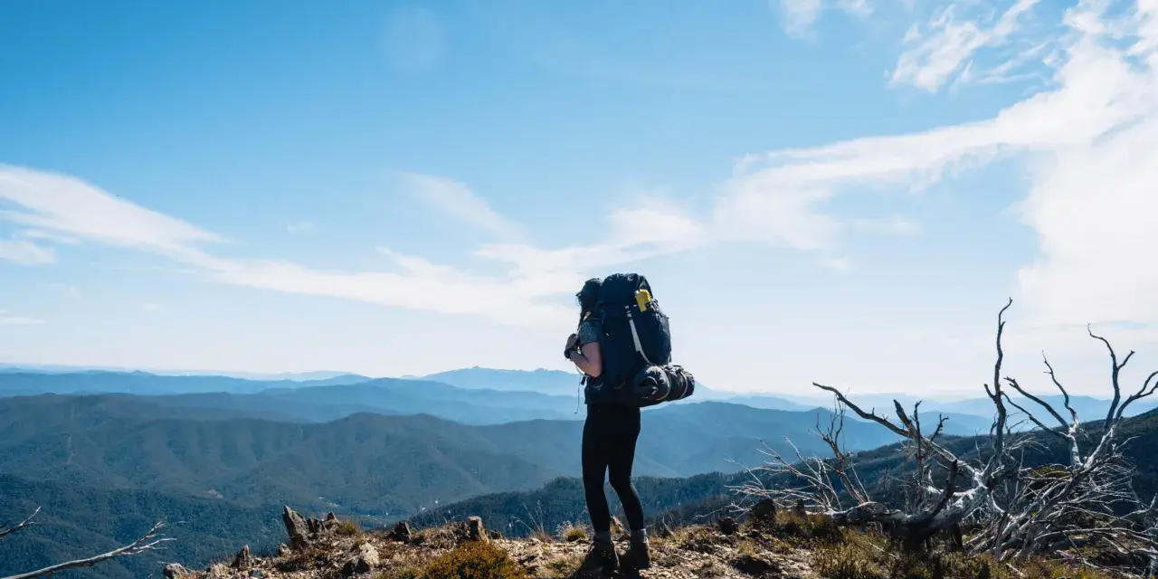 A lone hiker stands on a mountain ridge, ready for backpacking and hiking, with a vast expanse of blue mountains stretching to the horizon under a clear sky.