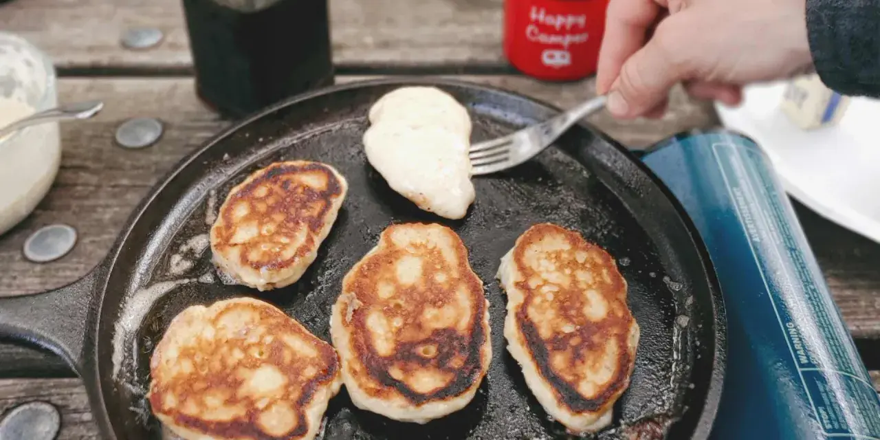 Frying pancakes in a cast iron skillet is a delicious answer to what to eat when camping. A fork lifts a fresh pancake, ready for syrup.