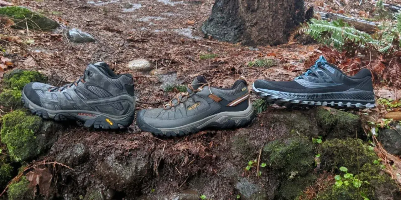 Three pairs of shoes on a wet forest floor: hiking boots, trail running shoes, and another pair of trail running shoes.