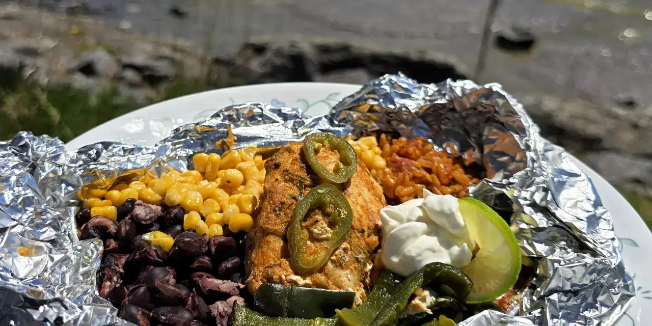 Delicious campfire chicken foil packets with corn, black beans, rice, jalapeños, and a lime wedge, ready to be enjoyed by the water.