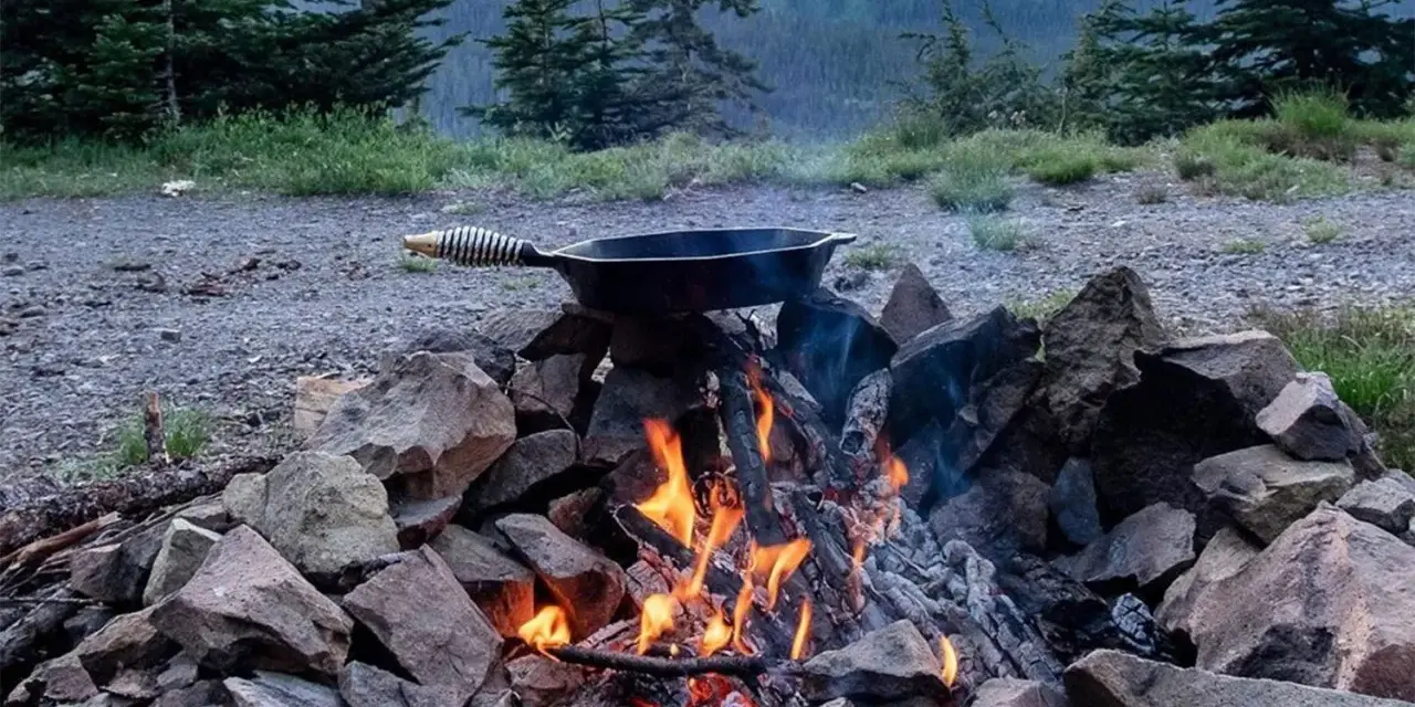 A cast iron skillet sits over a crackling campfire, ready for a delicious cast iron camping meal amidst a scenic mountain backdrop.