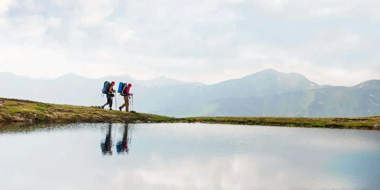 Two hikers with backpacks walk along a grassy ridge beside a calm lake, with misty mountains in the background. This image inspires thoughts on how to go backpacking.
