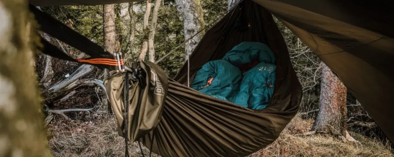 Cozy hammock camping setup in a forest. Two teal sleeping bags are nestled inside a brown hammock, ready for a night under the stars.