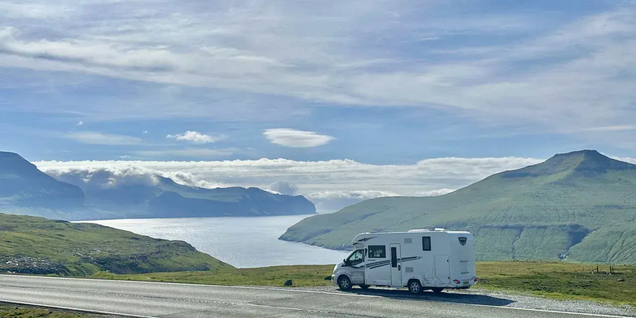 A white RV parked on a scenic road overlooking a bay and green mountains. This view is perfect for the best RV trip planner.