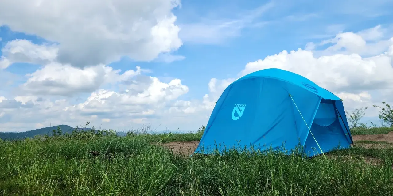 A bright blue Nemo tent stands in a grassy field under a cloudy sky. This Nemo Aurora tent is ready for adventure.