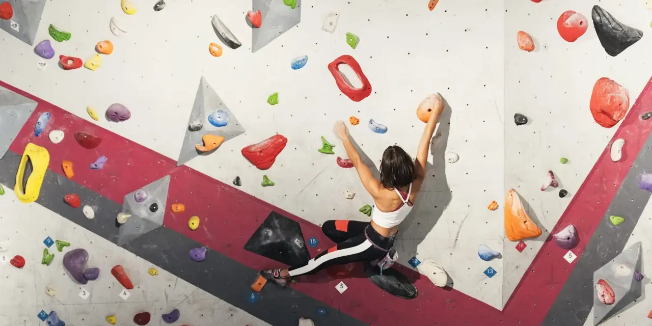 A woman is bouldering rock climbing, reaching for an orange hold on a colorful, textured wall.