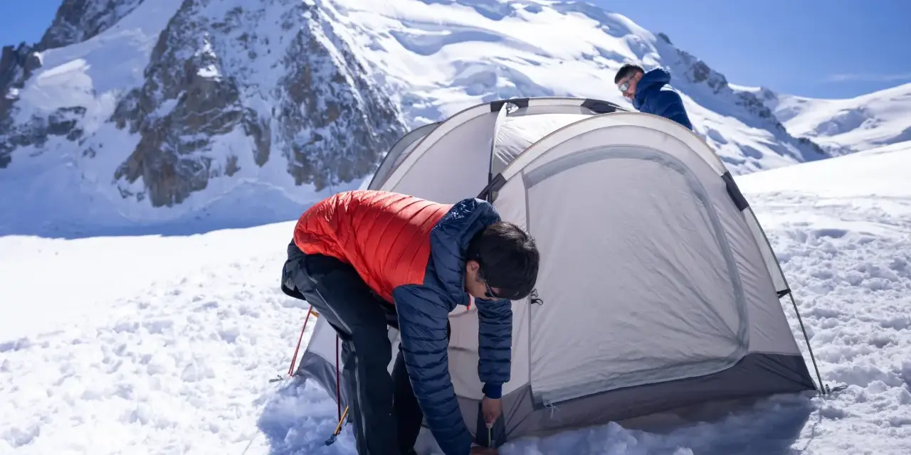 Two people set up a tent for winter camping amidst a snowy mountain landscape under a clear blue sky.